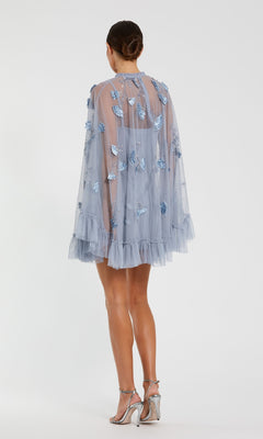 Woman wearing the Mac Duggal 9257 Long Sleeve Short Hoco Dress in sheer light blue with ruffles and butterfly appliqués, standing on a white background and paired with silver high-heeled sandals.