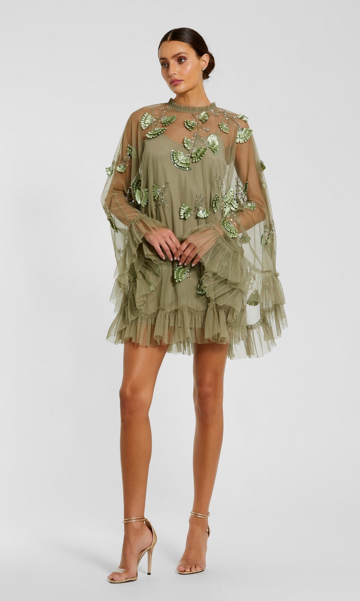A woman wears the Mac Duggal 9257 Long Sleeve Short Hoco Dress, a sheer olive green mini with leaf embroidery, ruffled hem, and flared sleeves, styled with nude high-heeled sandals.