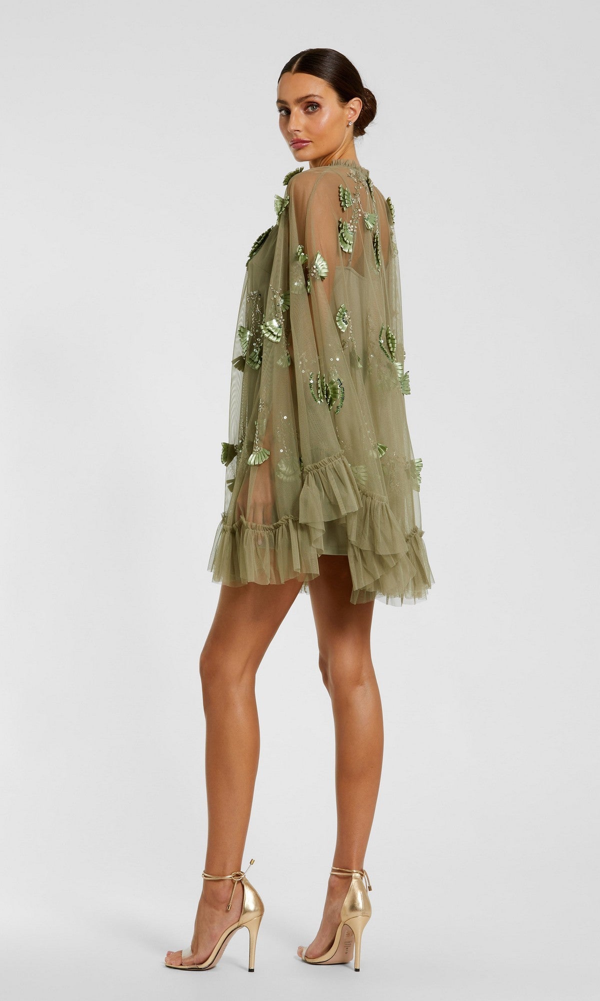 A woman in the Mac Duggal 9257 Long Sleeve Short Hoco Dress, a sheer olive green mini with sequin embellishments and ruffled hem and sleeves, wears gold heels against a plain background—perfect for homecoming.
