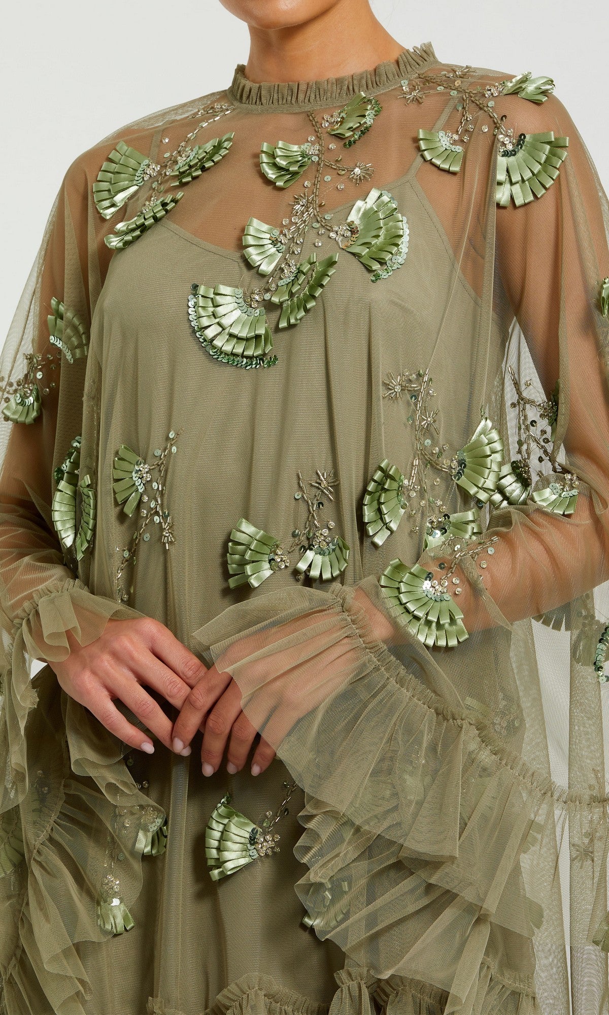 A person wears the Mac Duggal 9257 Long Sleeve Short Hoco Dress in olive green, featuring sheer fabric, shiny green applique fans, ruffles, and intricate beadwork.