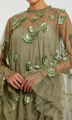 A person wears the Mac Duggal 9257 Long Sleeve Short Hoco Dress in olive green, featuring sheer fabric, shiny green applique fans, ruffles, and intricate beadwork.