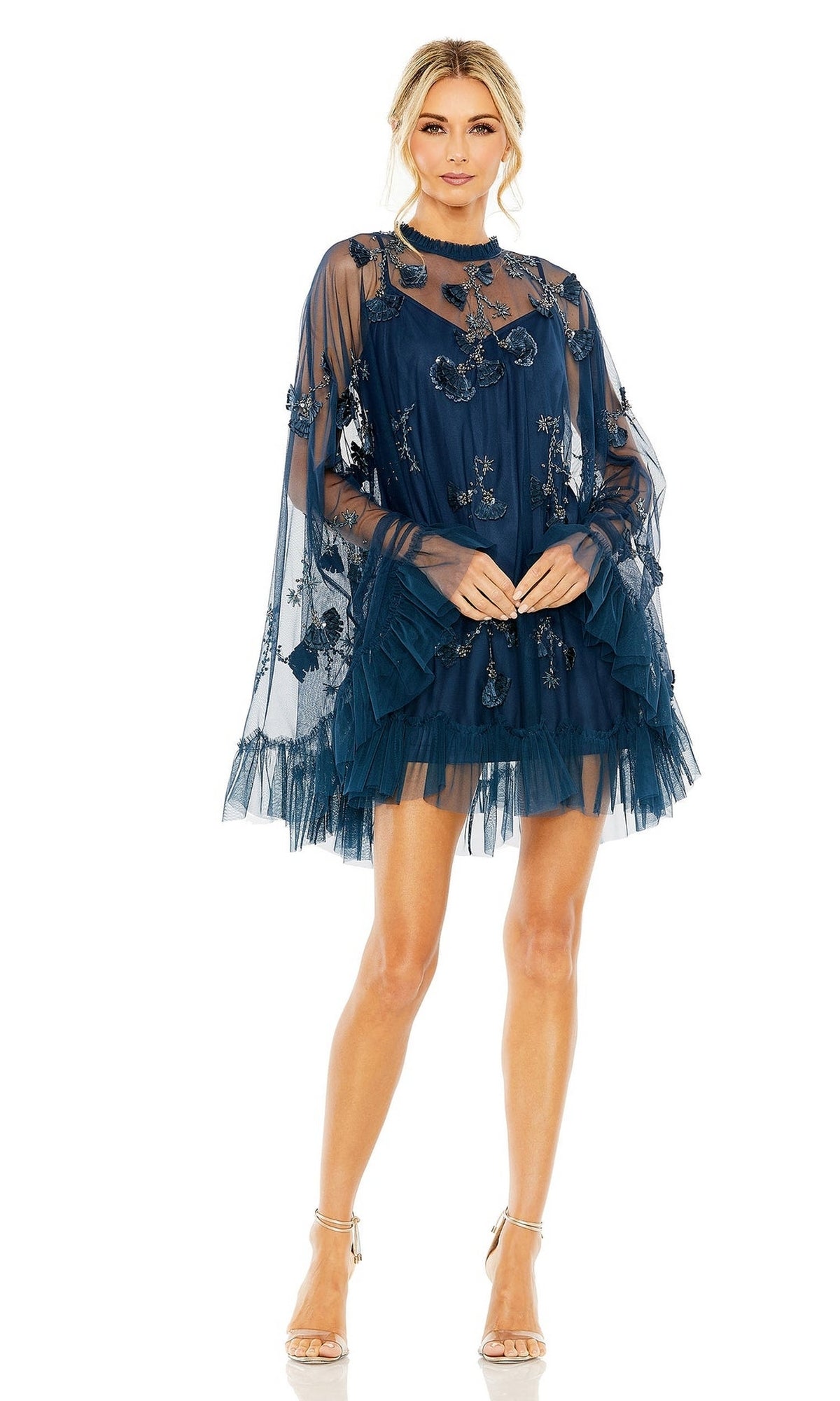 Woman wearing the Mac Duggal 9257 Long Sleeve Short Hoco Dress, featuring navy blue sheer embroidered fabric, floral patterns, tulle layers, and sheer cape sleeves, paired with strappy high-heeled sandals.