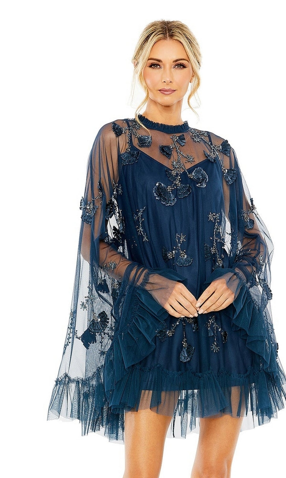 A woman poses against a white background in the elegant Mac Duggal 9257 Long Sleeve Short Hoco Dress, featuring a sheer navy blue design with floral appliqué, layered tulle, and ruffled accents.