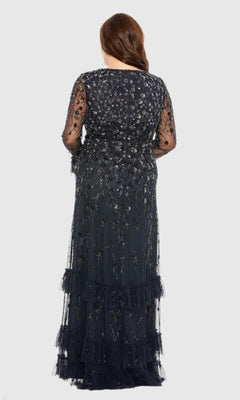 A woman stands facing away, wearing the Mac Duggal 9271 long plus-size formal dress in dark blue, featuring floor-length sequin beading, sheer beaded long sleeves, and intricate embroidery. Her hair is styled in loose waves down her back.