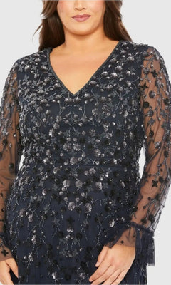 A person stands against a plain background wearing the Long Plus-Size Formal Dress: Mac Duggal 9271, featuring navy blue fabric, sheer sleeves, and intricate silver floral embroidery.