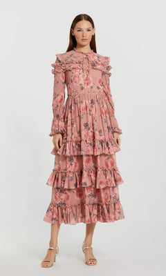 A woman wearing the Midi Wedding Guest Dress: Mac Duggal 9306 in Rose Multi, a long-sleeved chiffon dress featuring pink floral patterns, ruffles, pleats, and a tiered skirt, stands against a plain white background.