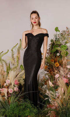 A woman stands among lush greenery and colorful flowers in the Off-Shoulder Long Glitter Prom Dress: Eureka 9306, the fitted black gown making an elegant statement against a plain background.