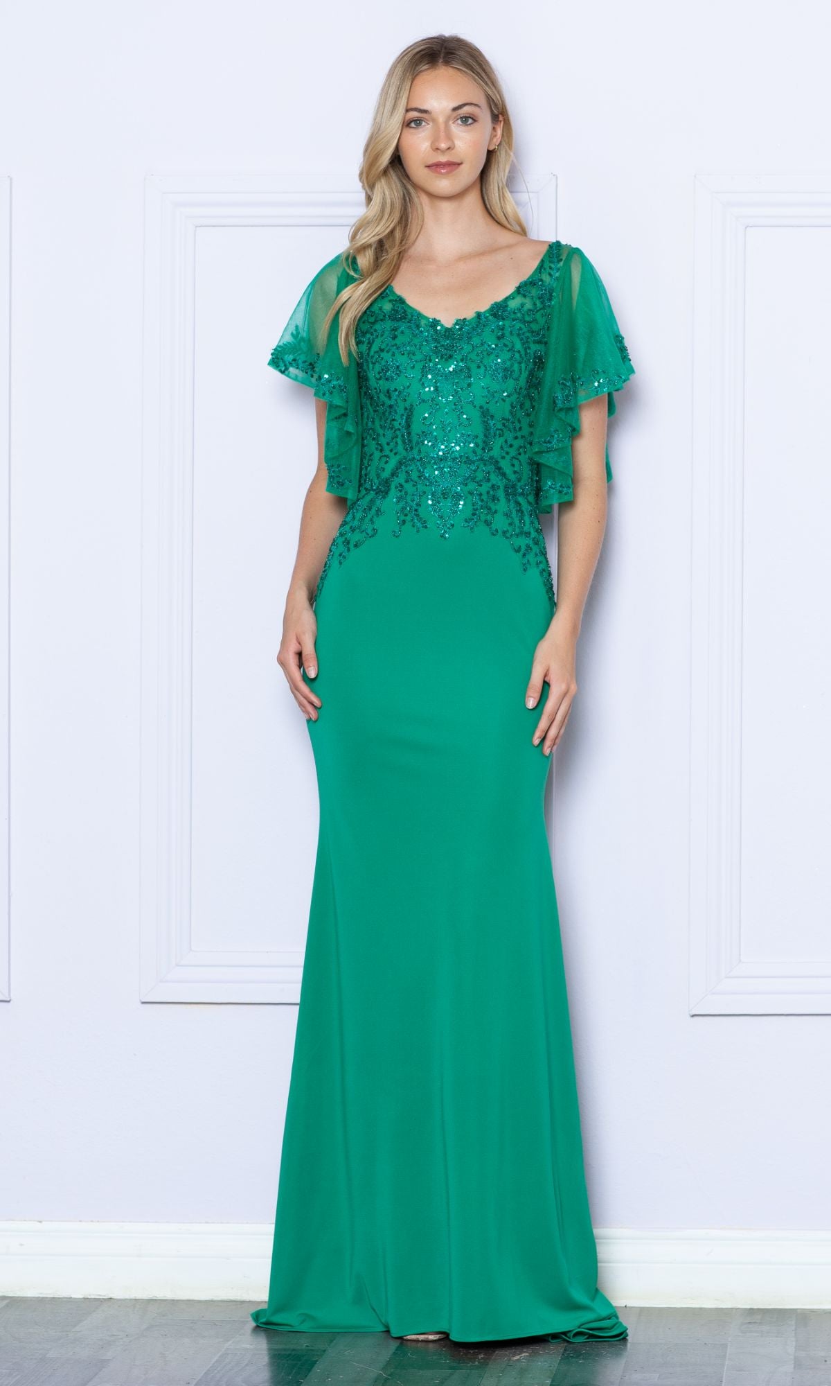 A woman stands against a light-colored wall wearing the Flutter-Sleeve Embellished Long Formal Dress 9318, a plus-size green gown with sequin details and short, sheer flutter sleeves.