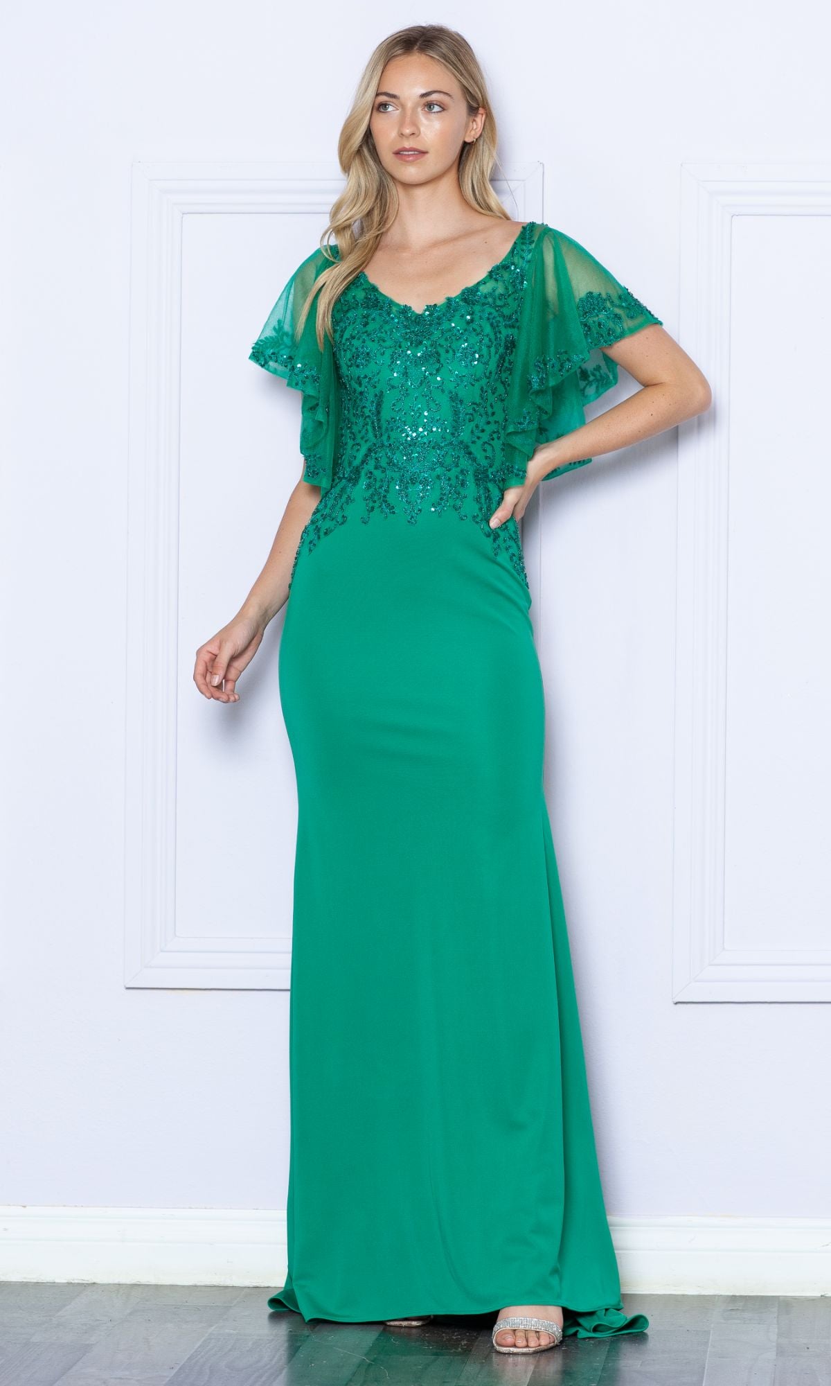 A woman stands against a light-colored wall wearing the Flutter-Sleeve Embellished Long Formal Dress 9318 in emerald, featuring sequin details and sheer flutter sleeves.