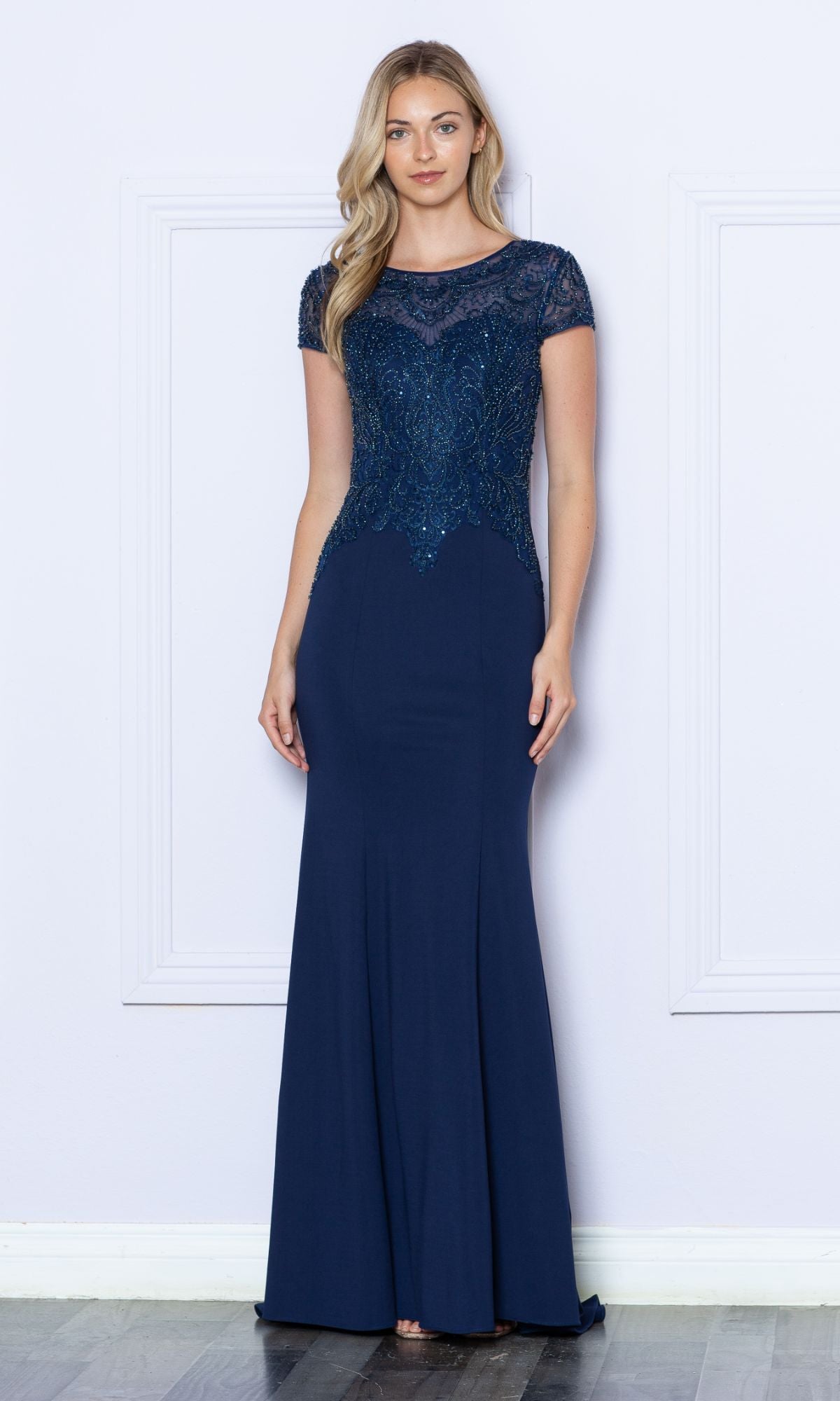 A woman stands indoors wearing the Beaded Short Sleeve Bodice Long Formal Dress 9320 in navy, featuring short sleeves and a bodice intricately embellished with sparkling beads.