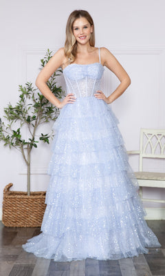 A woman stands indoors wearing the Corset-Back Long Tiered Sequin Prom Ball Gown 9328, a sparkly light blue ball gown with spaghetti straps and delicate tiers. A potted plant and a white bench are in the background.