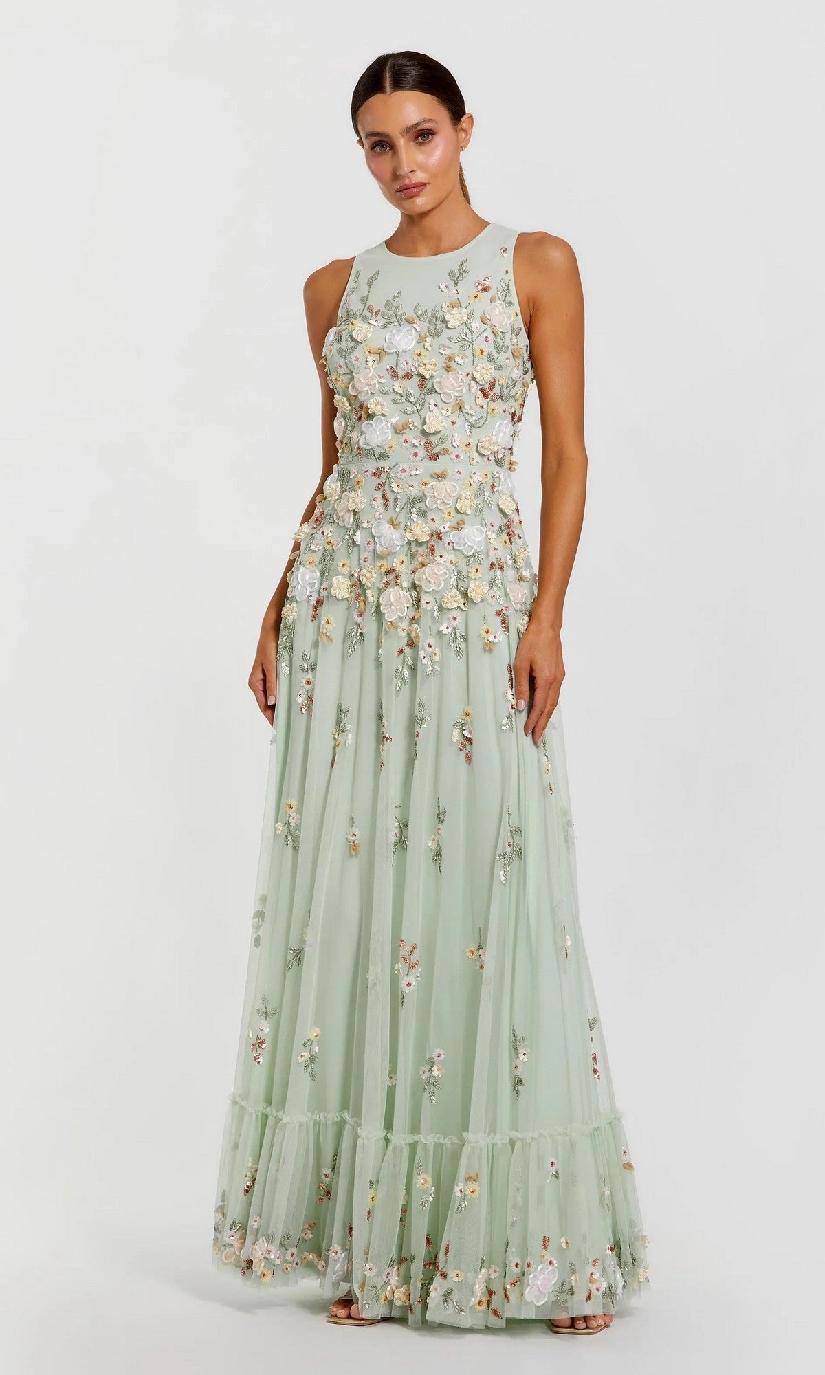 A woman wearing the Mac Duggal 9334 long formal dress in sage multi, featuring a sleeveless, floor-length design with floral embroidered appliqué details, stands against a plain light background.