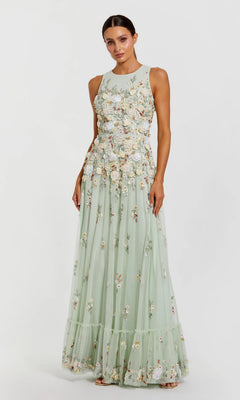 A woman wearing the Mac Duggal 9334 long formal dress in sage multi, featuring a sleeveless, floor-length design with floral embroidered appliqué details, stands against a plain light background.
