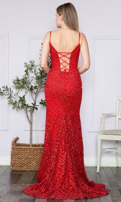 A woman stands facing away in a Glitter-Lace Long Corset Prom Dress 9354, its fitted red silhouette and lace-up back highlighted beside a potted plant and white chair in a softly lit room.