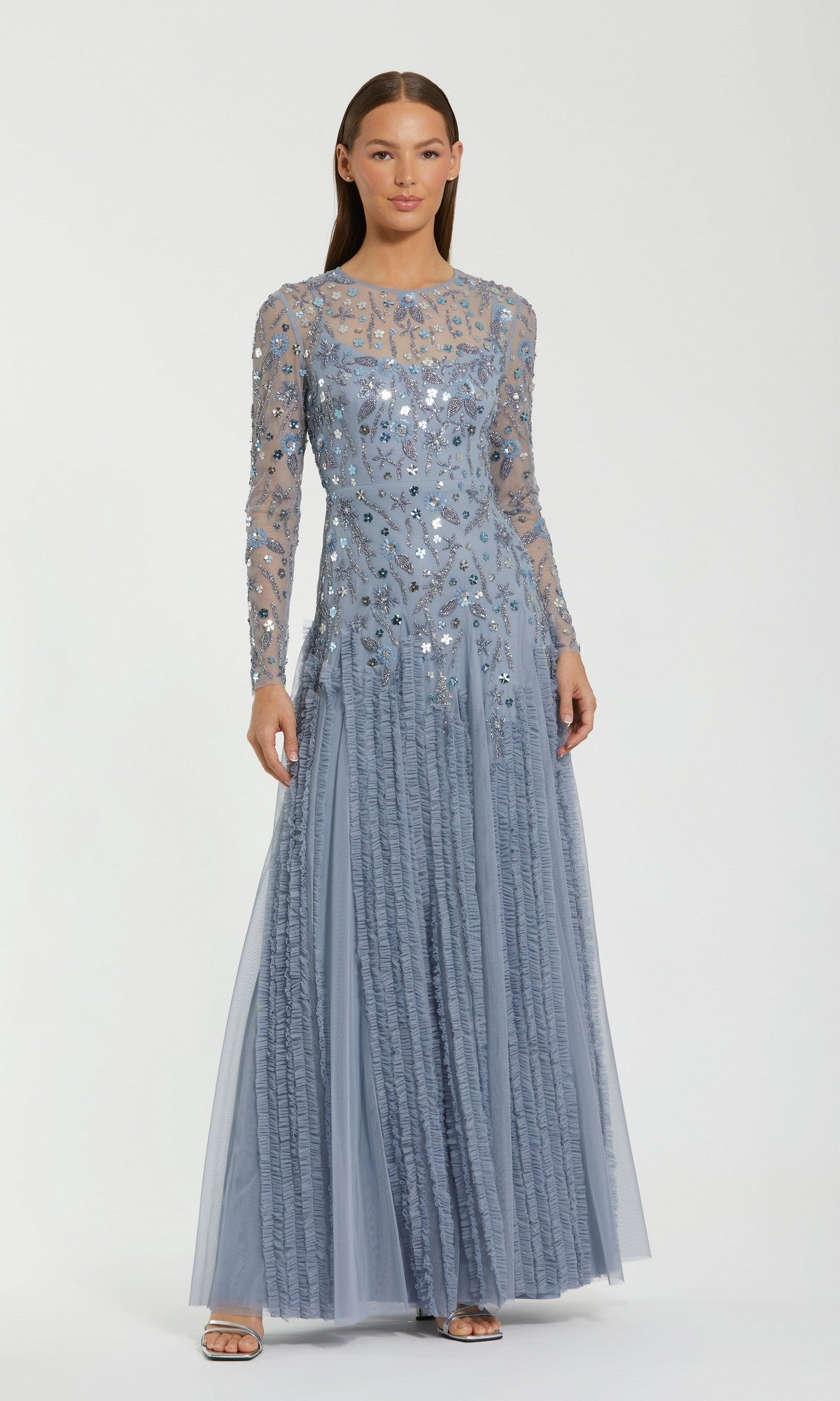 A woman stands wearing the Mac Duggal 9357 long formal dress in slate blue, featuring sheer long sleeves and silver sequin embellishments, posing against a plain white background.