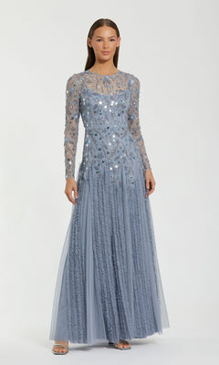 A woman stands wearing the Mac Duggal 9357 long formal dress in slate blue, featuring sheer long sleeves and silver sequin embellishments, posing against a plain white background.
