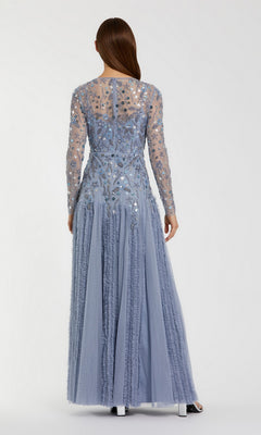 A woman stands facing away, wearing the Mac Duggal 9357 Long Formal Dress in light blue—a floor-length, long-sleeved gown crafted from sheer, embellished fabric with delicate ruffled skirt details.