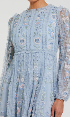 A person wearing the Mac Duggal 9365 short wedding guest dress, featuring light blue floral embroidery, sheer long sleeves, and ruffle details.