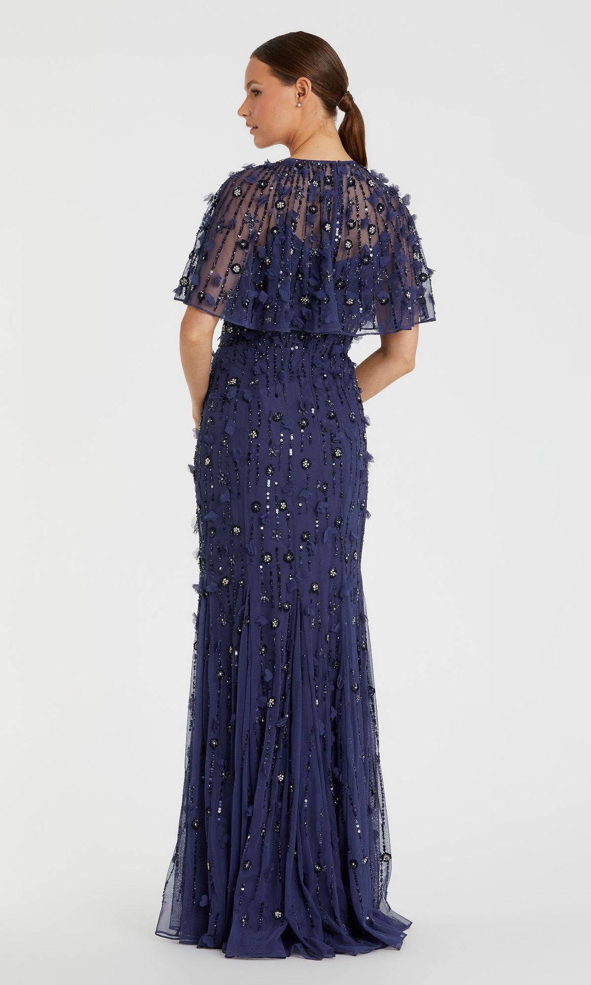A woman faces away in the Mac Duggal 93653 Beaded-Cape Long Formal Dress, a dark blue gown with a sheer, embellished overlay and sequin details. Her hair is styled in a low ponytail.
