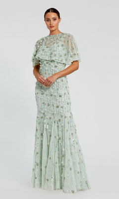 A woman wears the Mac Duggal 93653 Beaded-Cape Long Formal Dress in pale green, featuring a high neck, sequins, and a sheer capelet—an elegant mother-of-the-bride look—against a plain background.