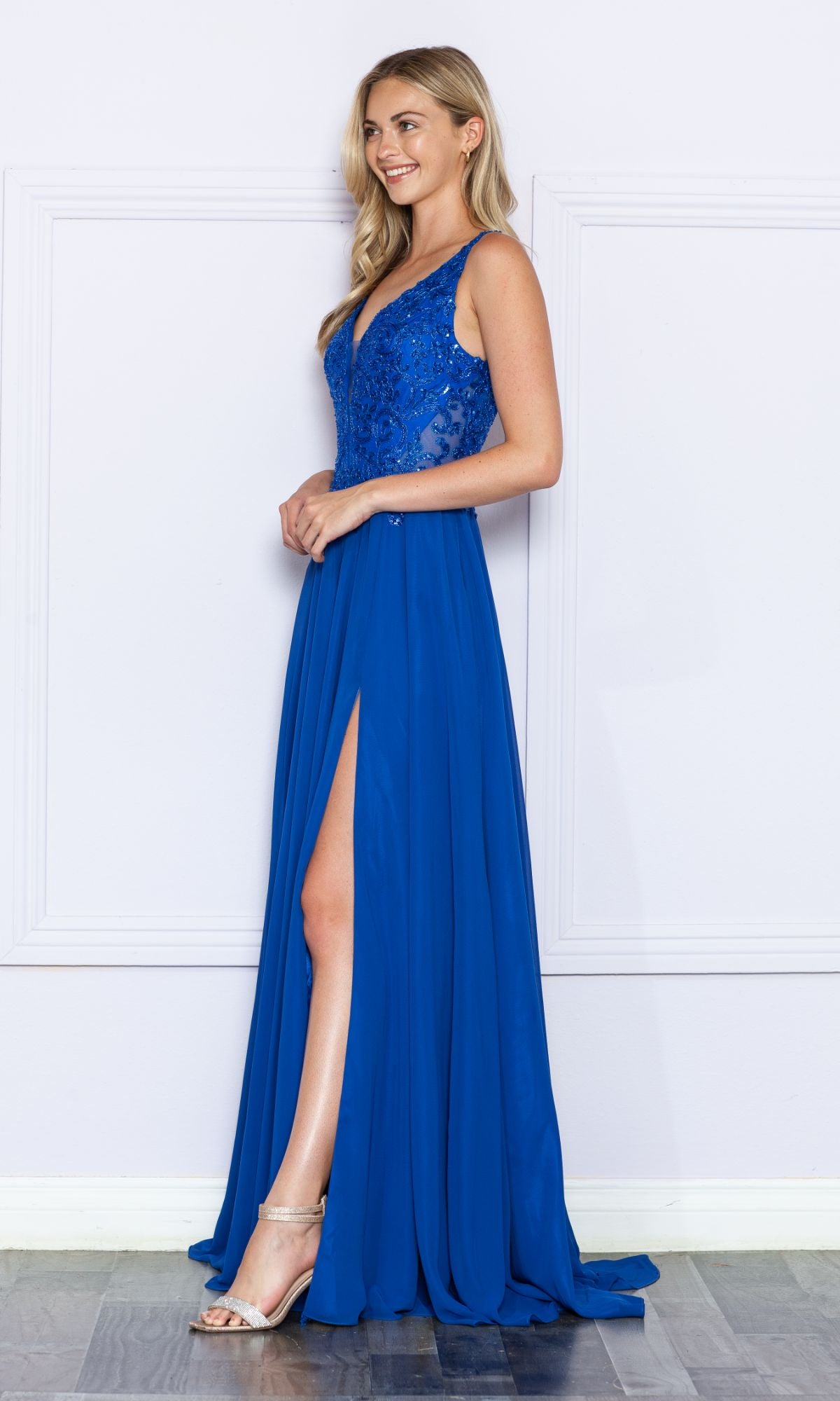 A woman stands sideways in the Beaded-Bodice Long A-Line Prom Dress 9366, a sleeveless royal blue gown with a sparkly beaded bodice, thigh-high slit, and paired with silver high-heeled sandals.