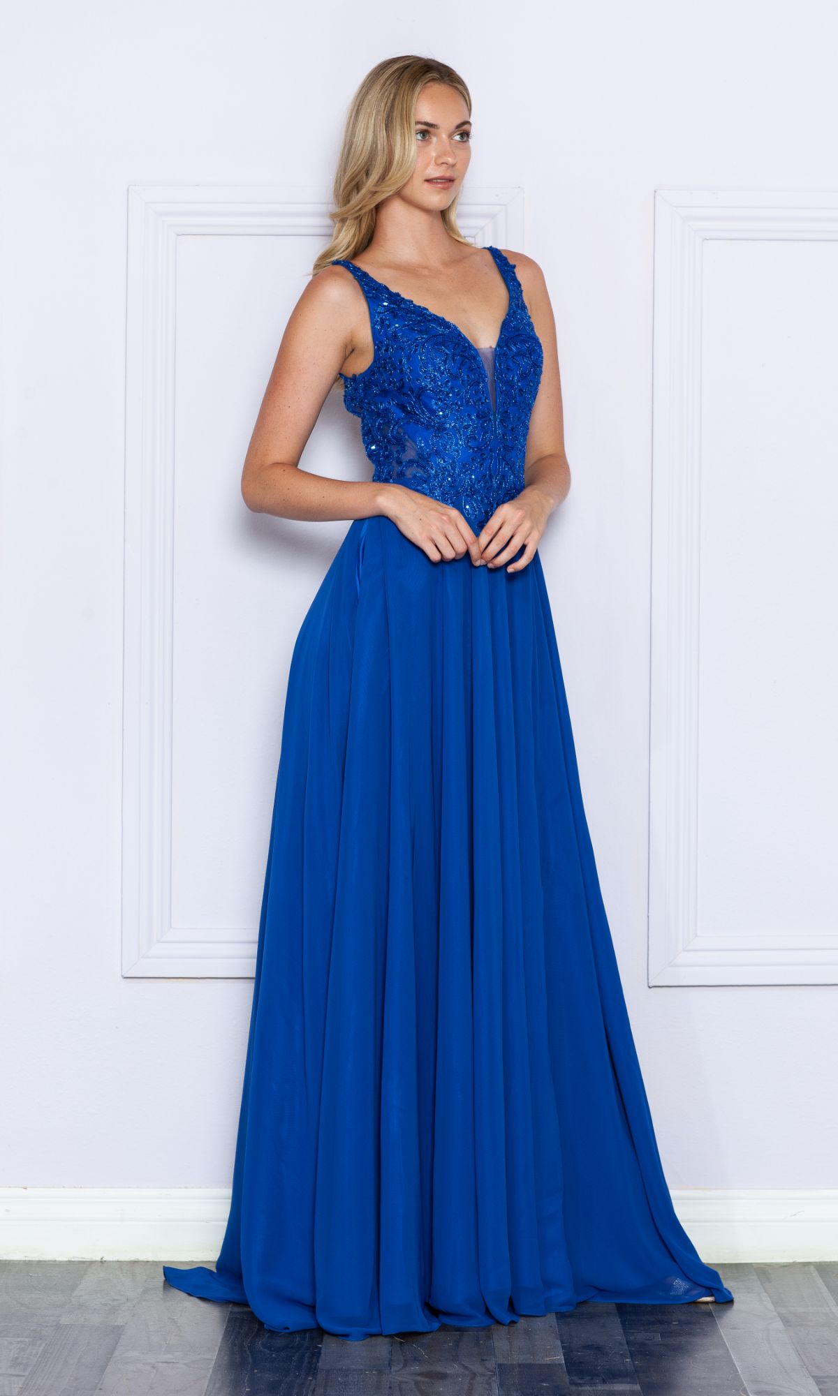 A woman stands against a pale wall wearing the Beaded-Bodice Long A-Line Prom Dress 9366 in royal blue, featuring a sparkly bodice and a flowing floor-length skirt.
