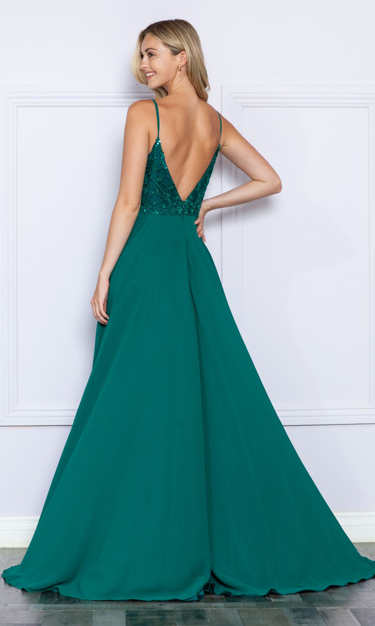 A woman in the Sequin-Bodice Long A-Line Prom Dress 9368—emerald green, sleeveless with a deep V-back, fitted bodice, and flowing skirt—stands against a white wall.
