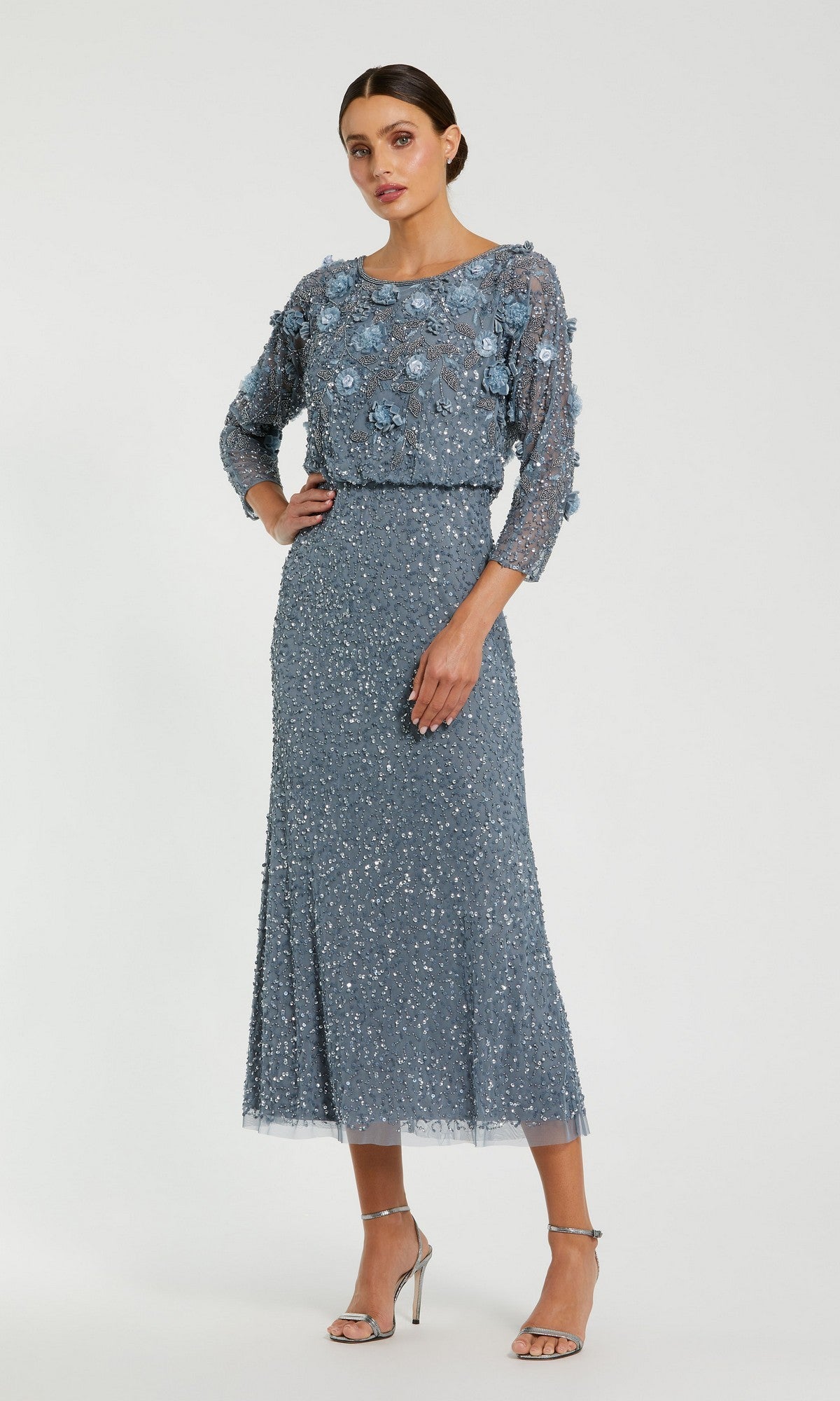 A woman stands against a plain background wearing the Mac Duggal 9373 Tea-Length Wedding Guest Dress in slate blue, featuring three-quarter sleeves and an elegant beaded silhouette adorned with stunning floral and sequin embellishments, paired with silver high-heeled sandals.