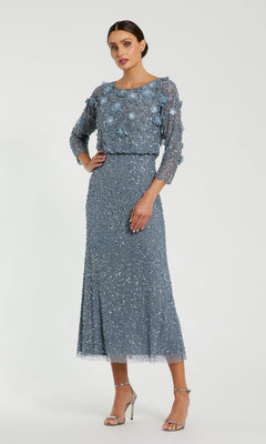 A woman stands against a plain background wearing the Mac Duggal 9373 Tea-Length Wedding Guest Dress in slate blue, featuring three-quarter sleeves and an elegant beaded silhouette adorned with stunning floral and sequin embellishments, paired with silver high-heeled sandals.