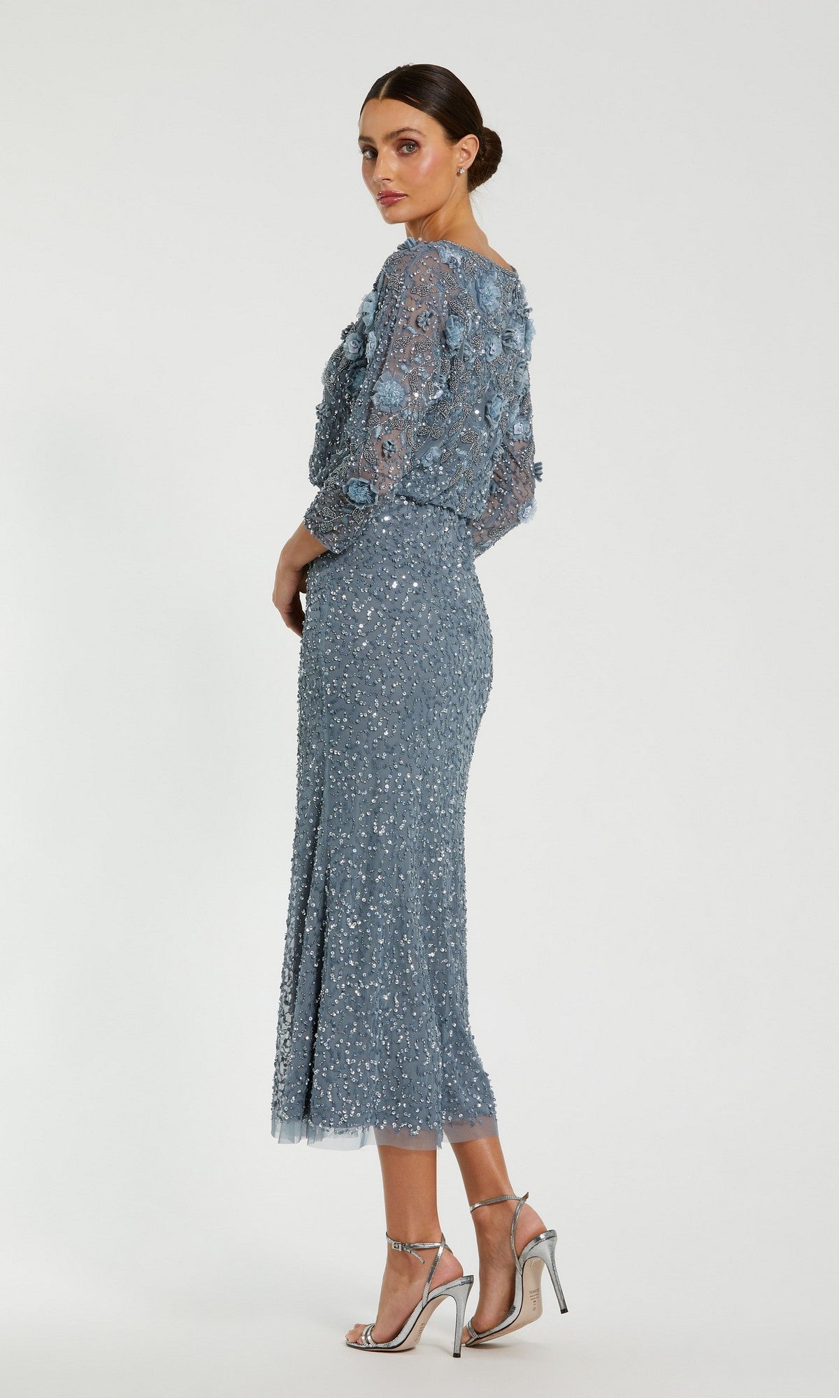 A woman wearing the Mac Duggal 9373 tea-length wedding guest dress—featuring blue sequin embellishments, sheer sleeves, and a graceful beaded silhouette—stands against a plain white background, paired with silver high-heeled sandals.