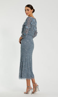A woman wearing the Mac Duggal 9373 tea-length wedding guest dress—featuring blue sequin embellishments, sheer sleeves, and a graceful beaded silhouette—stands against a plain white background, paired with silver high-heeled sandals.