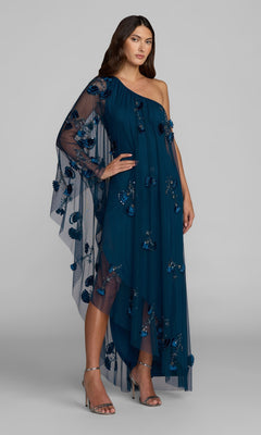 A woman stands against a plain gray background, wearing the Mac Duggal 9374 Long Formal Dress in twilight—a one-shoulder gown featuring floral appliqués, a sheer cape sleeve, and paired with silver high-heeled sandals for an elegant look.