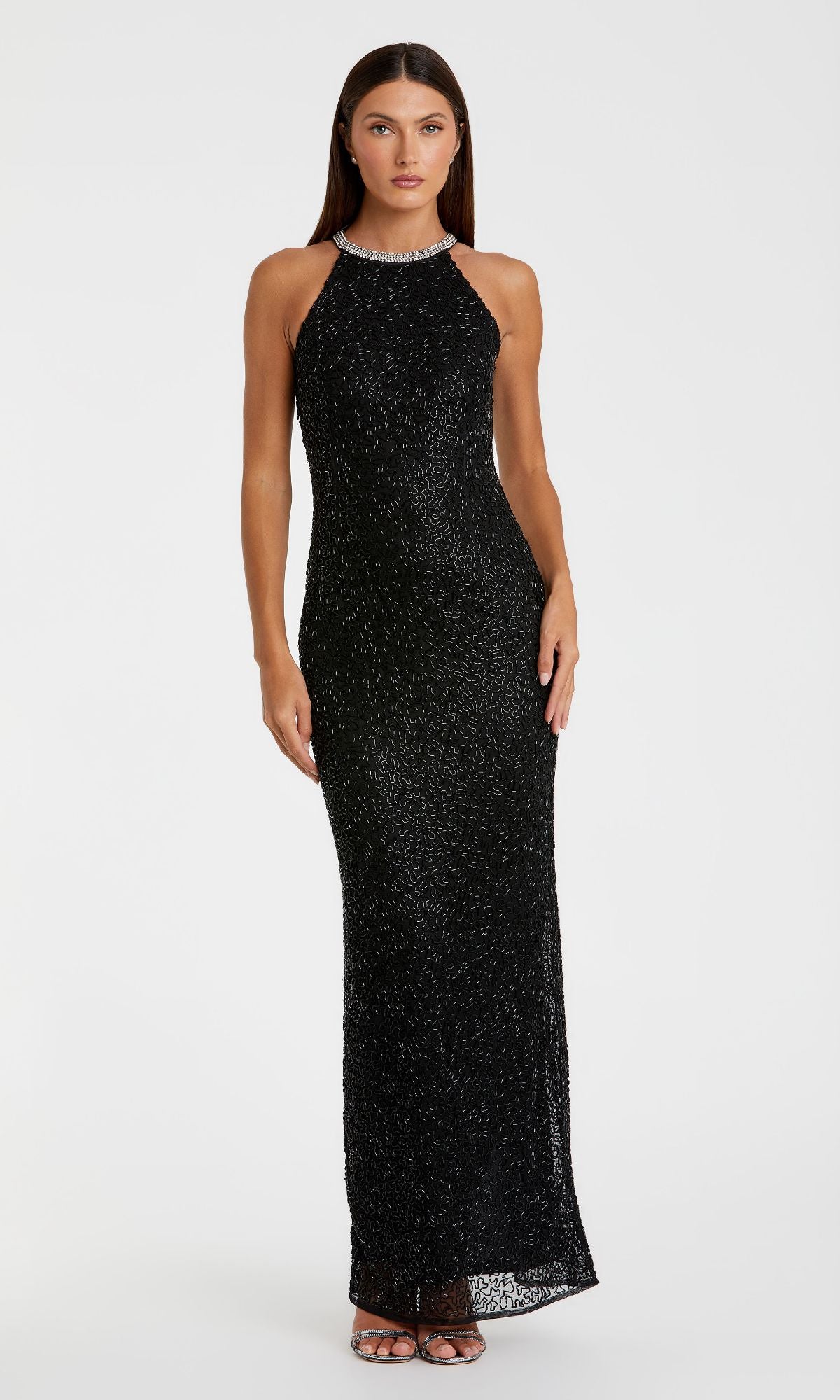 A woman faces forward in the Mac Duggal 93742 long black beaded formal dress, featuring a sleeveless design, high neckline, and paired with silver strappy heels against a plain white background.