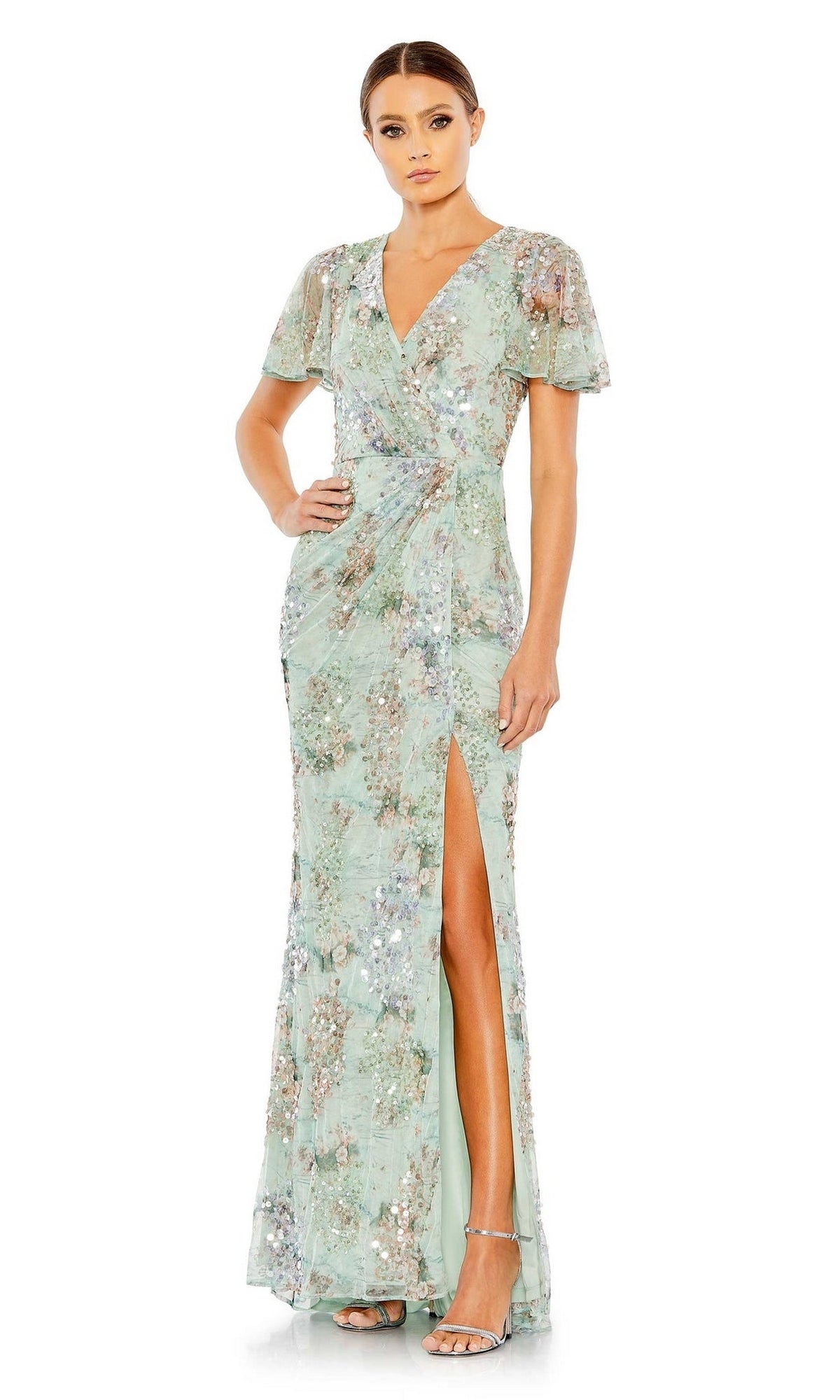 A woman poses with one hand on her hip, wearing the Mac Duggal 93749 Short Sleeve Beaded Formal Dress in light green, featuring floral patterns, a V-neckline, and a thigh-high slit.