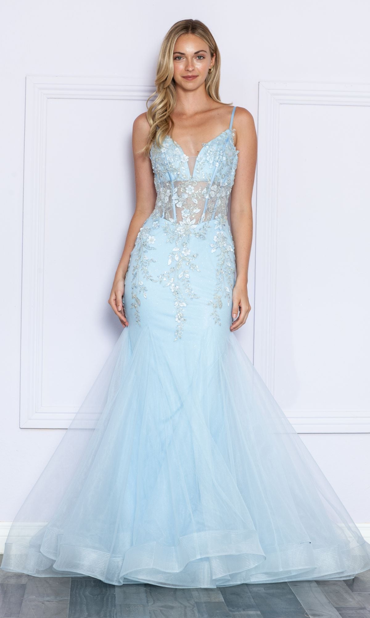 A woman poses against a light-colored wall in the Sheer-Corset-Bodice Long Mermaid Prom Dress 9374, a light blue, floor-length gown featuring sheer panels and silver floral embroidery.