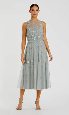 A woman stands against a plain background wearing the Midi Party Dress: Mac Duggal 9380 in sage—a sleeveless party dress adorned with beaded floral embroidery and a sheer overlay—paired with silver high-heeled sandals.