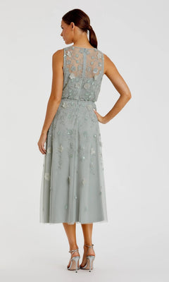 A woman stands facing away, wearing the Mac Duggal 9380 Midi Party Dress—a sleeveless sage-green party dress with beaded floral embroidery and sheer detailing—paired with silver high-heeled sandals.