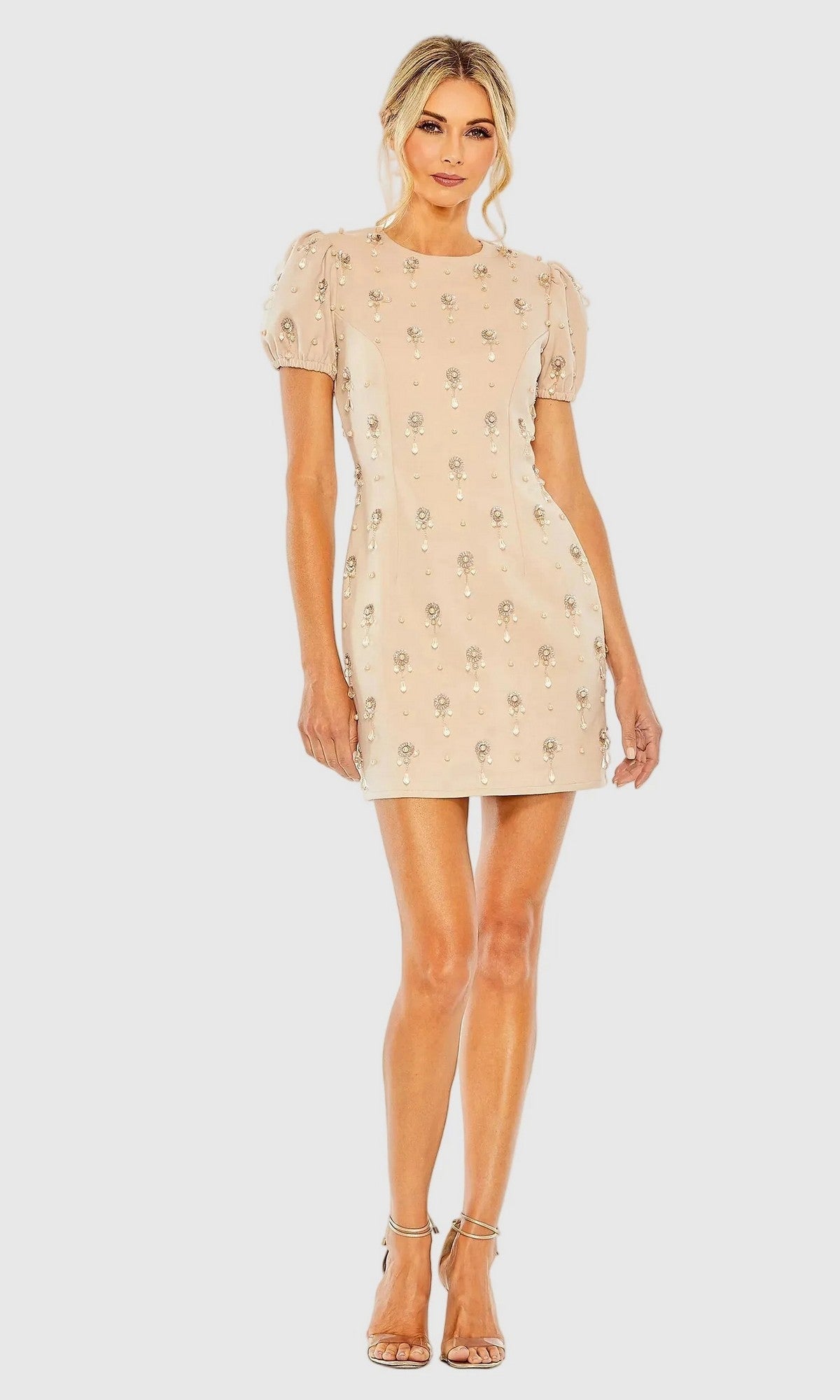A woman stands against a plain light background wearing the Mac Duggal 93836 Short Party Dress—a nude pink mini dress featuring puff sleeves and delicate floral embellishments—paired with strappy heels for an elegant, feminine look.