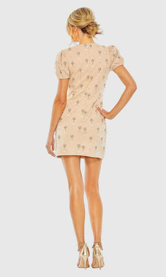 A woman stands facing away against a plain background, wearing the Mac Duggal 93836 Short Party Dress—a beaded mini dress with floral embellishments—paired with gold high-heeled sandals.