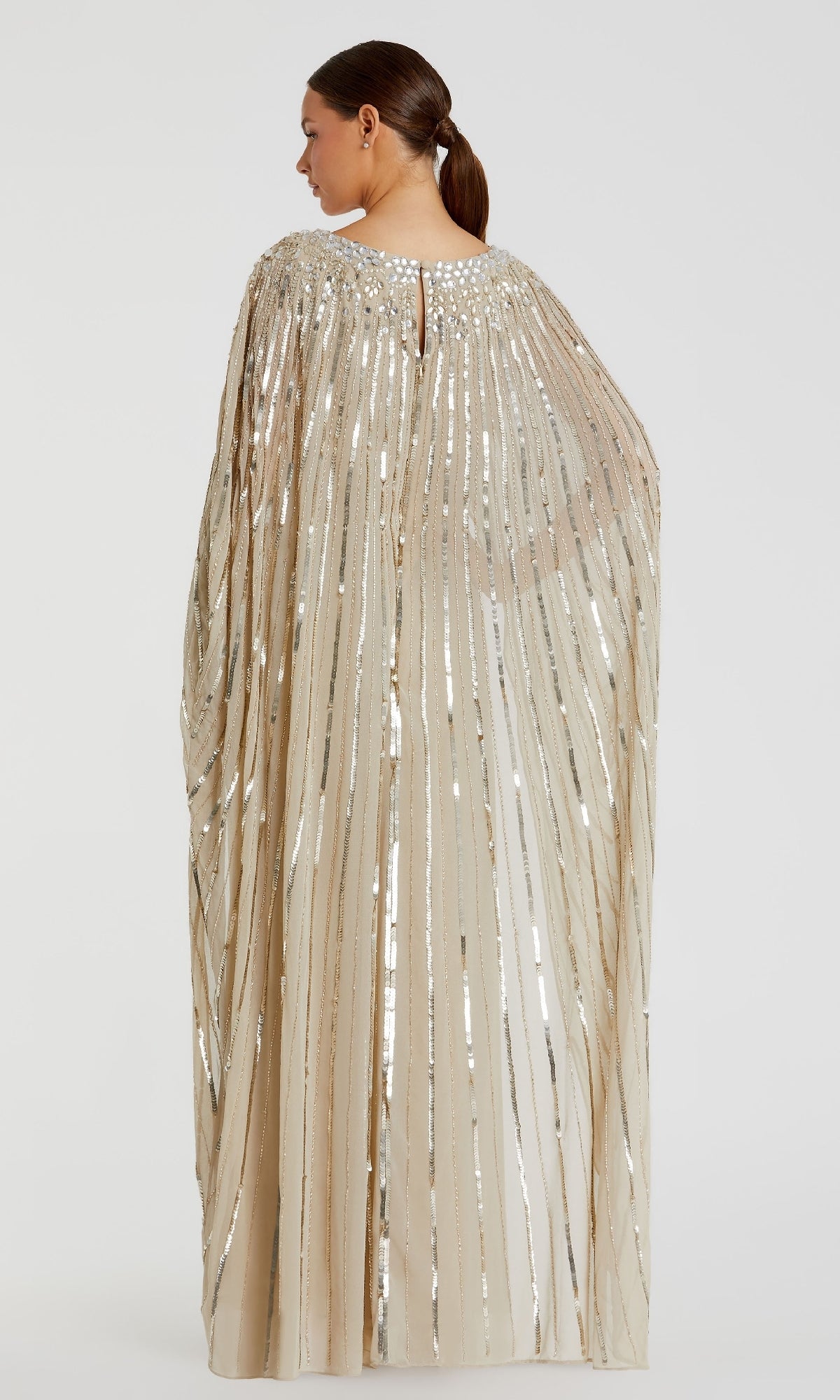 A woman faces away, wearing the Mac Duggal 93869 Caped Long Sequin Formal Dress—a beige gown with silver sequin vertical stripes and a cape-like design—standing against a plain light background.