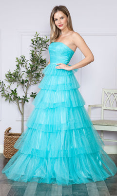 A woman stands indoors wearing the Strapless Long Tiered Glitter Prom Ball Gown 9386 in turquoise tulle; tiered ruffles cascade down, with a potted plant and white chair in the background.