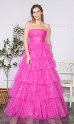 A woman stands indoors wearing the Strapless Long Tiered Glitter Prom Ball Gown 9386 in bright pink tulle with multiple ruffled layers, set against a simple background featuring a plant and a white chair.