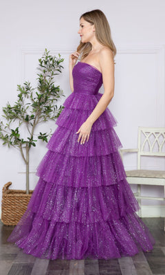 A woman stands indoors wearing the Strapless Long Tiered Glitter Prom Ball Gown 9386—a floor-length purple tulle dress. A potted plant and a white chair are in the background.