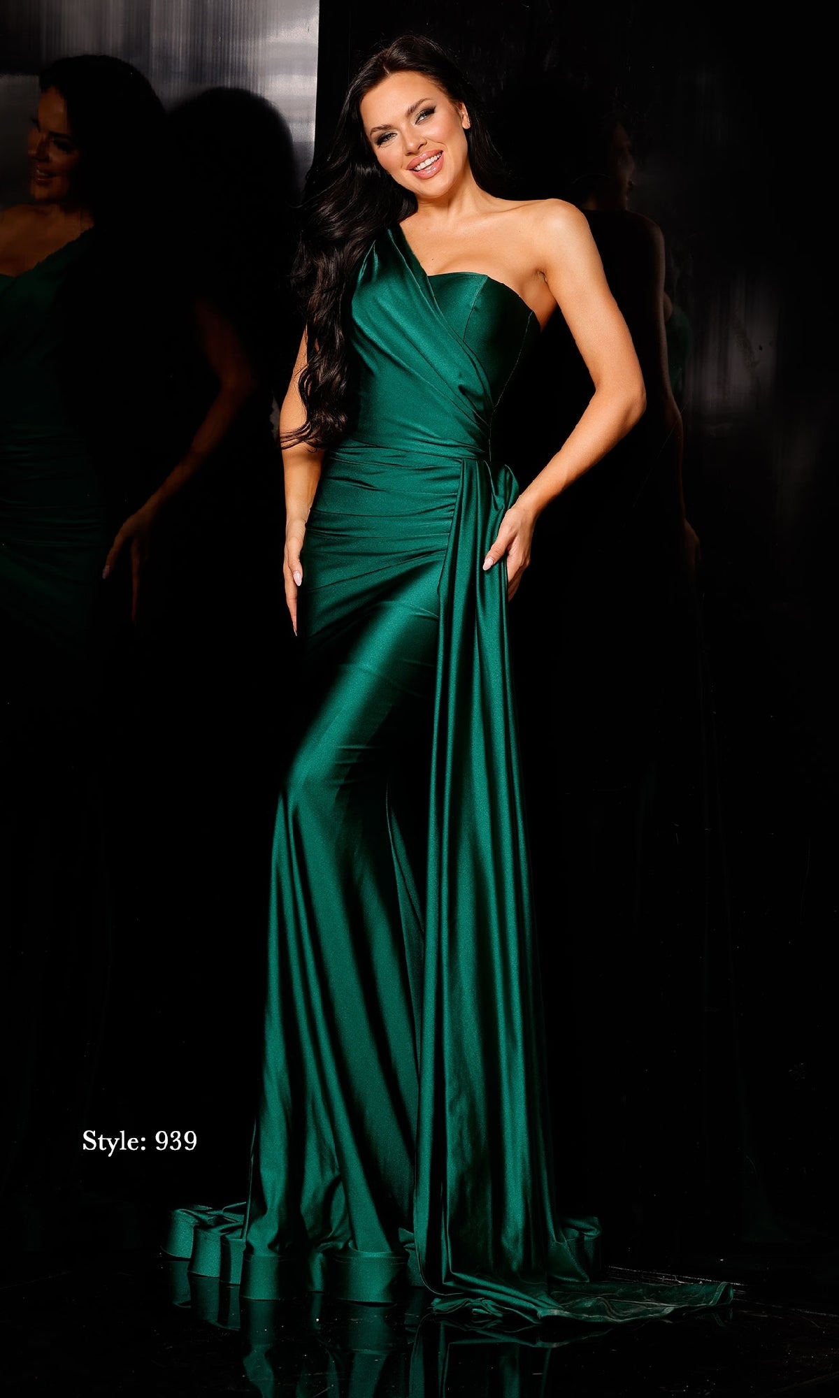 A woman models the Side-Drape Long Prom Dress: Jessica Angel 939 in emerald green, featuring a shiny one-shoulder design with a fitted bodice, draped sash, and flowing mermaid skirt against a dark background.