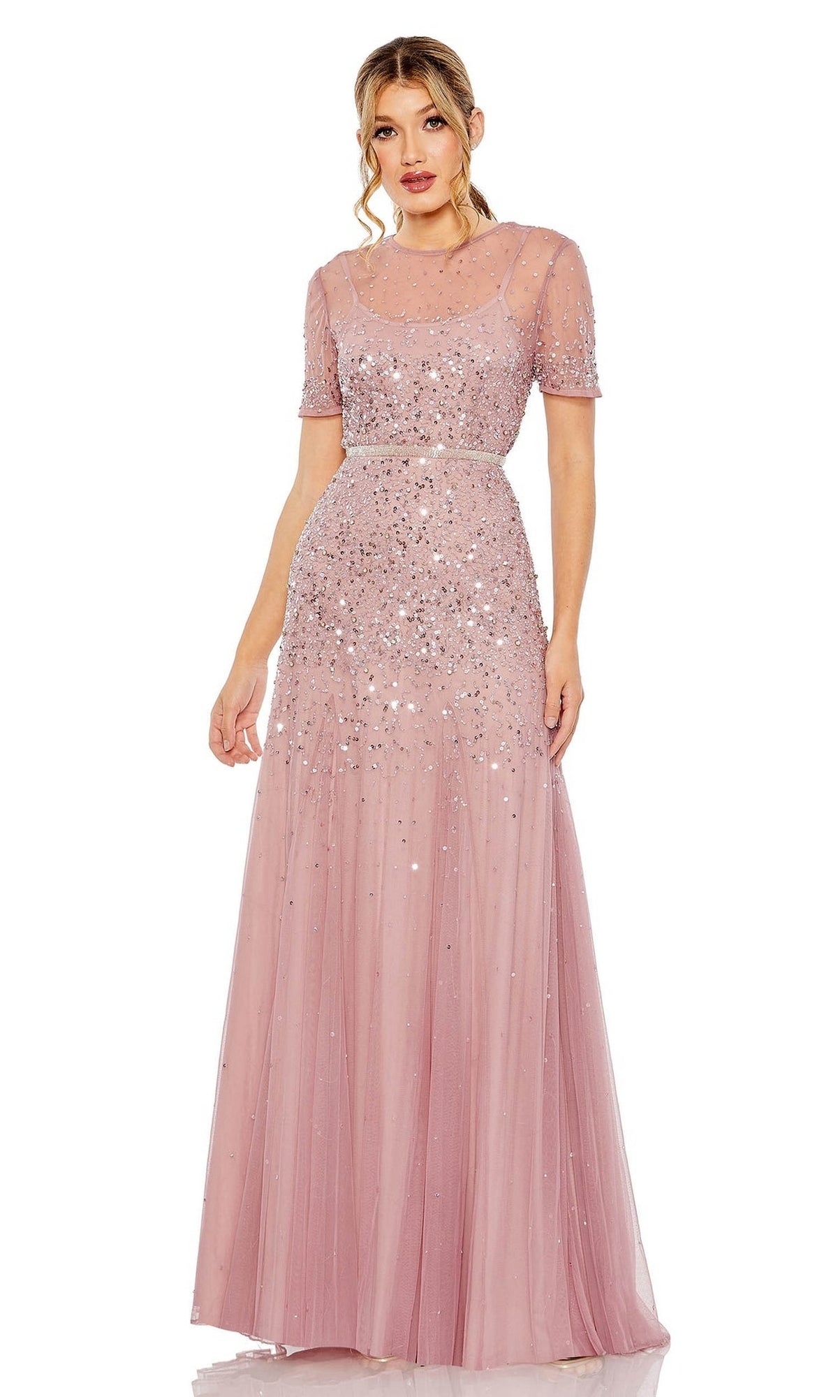 A woman stands against a white background wearing the Mac Duggal Sheer High-Neck Sequin Formal Dress 93909—a floor-length, pink gown with short sleeves, a beaded waist, and sheer overlay.