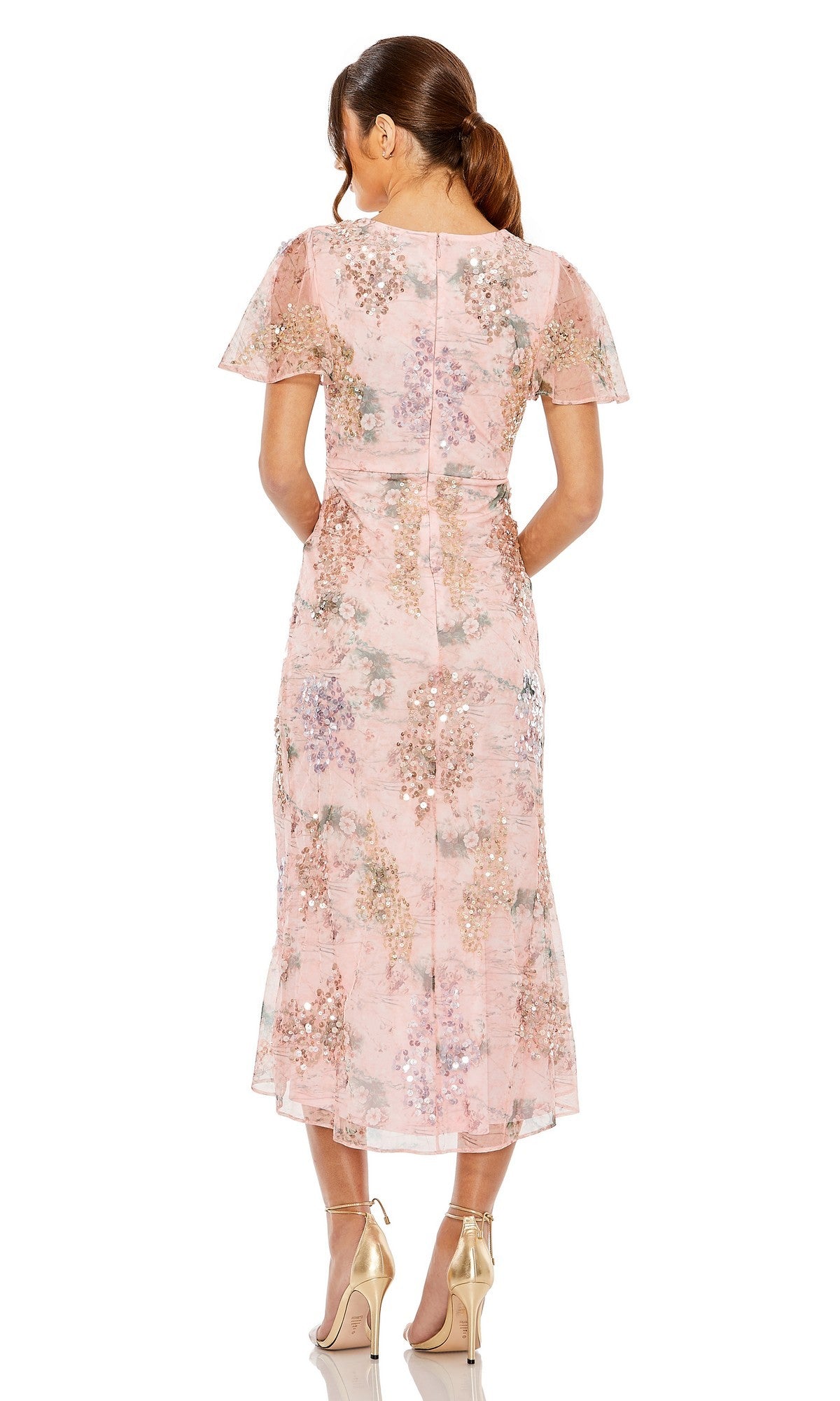 A woman stands facing away on a plain white background, wearing the Mac Duggal 93949 Midi Wedding Guest Dress, a pink floral wrap dress with short sleeves, paired with gold high heels.