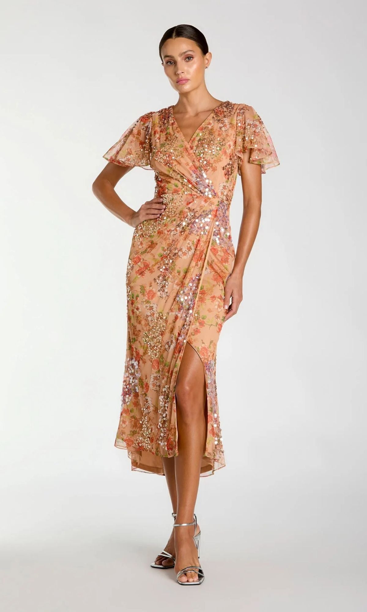 A woman wears the Mac Duggal 93949 Midi Wedding Guest Dress in apricot, featuring a floral print, faux-wrap design, short flutter sleeves, v-neckline, sequins, and a front slit. She completes the look with silver strappy heels.