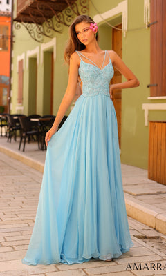 A woman in the Amarra Open-Back Long A-Line Prom Dress 94008, featuring a light blue beaded bodice and spaghetti straps, stands on a cobblestone street, gracefully holding one side of her floor-length gown.