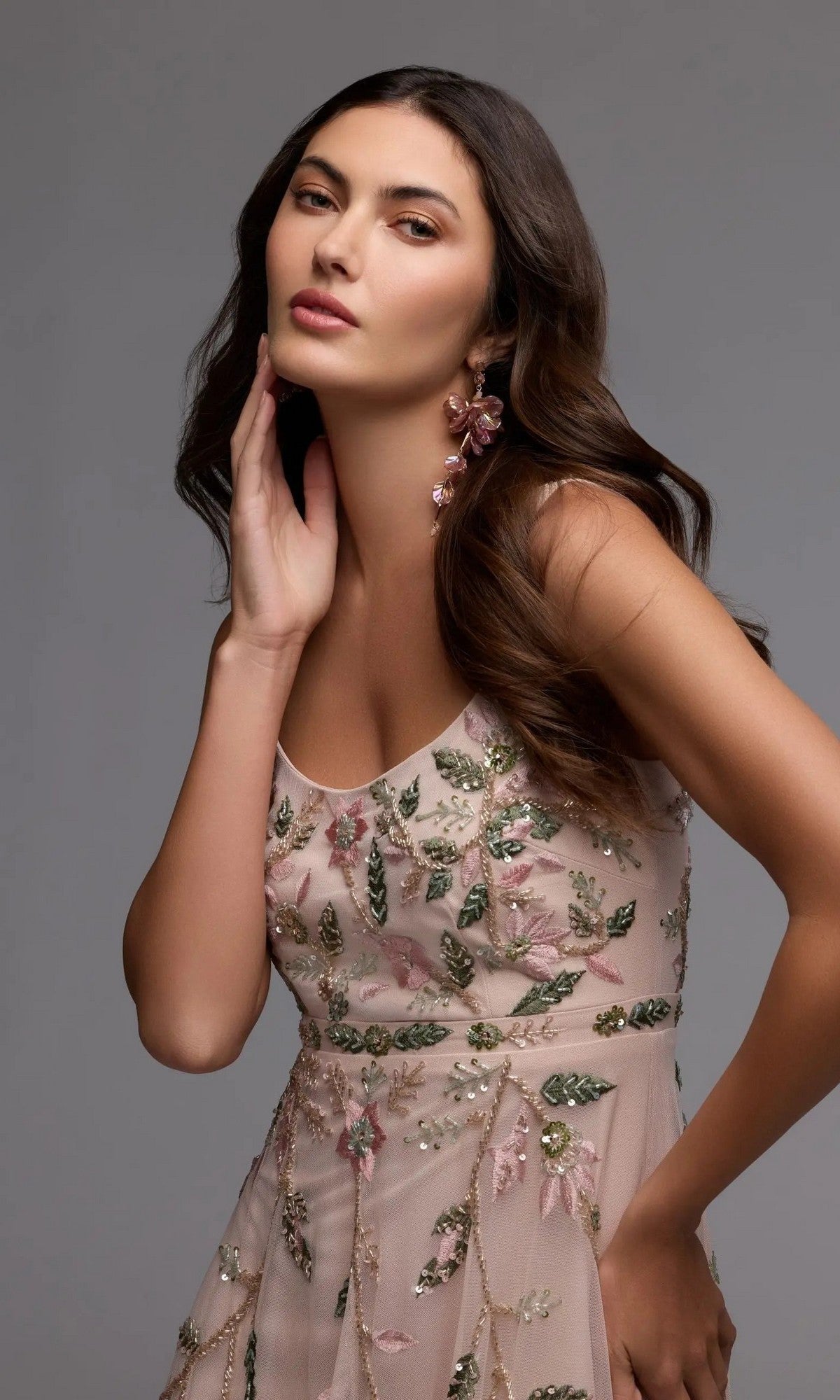 A woman with long brown hair poses with one hand touching her face against a plain gray background, wearing the Mac Duggal 9402 long formal dress—a sheer party gown adorned with floral beaded embroidery.