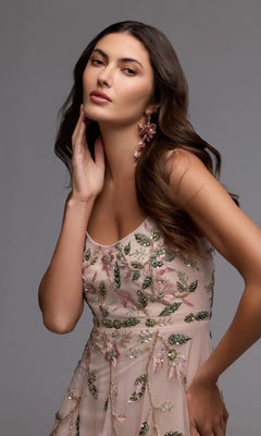 A woman with long brown hair poses with one hand touching her face against a plain gray background, wearing the Mac Duggal 9402 long formal dress—a sheer party gown adorned with floral beaded embroidery.